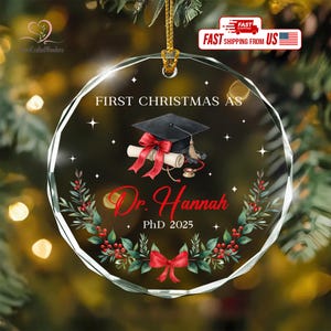 May include: Clear, round Christmas ornament with a graduation theme. The ornament features the text "FIRST CHRISTMAS AS Dr. Hannah PhD 2025" and a graduation cap, scroll, and red bow design. A festive holiday decoration.