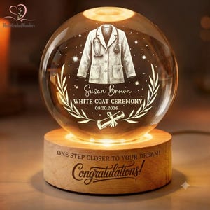 May include: A spherical glass night light on a wooden base. The glass features an etched white coat, a stethoscope, a diploma, and the text: Susan Brown, WHITE COAT CEREMONY, 09.20.2026. The base is engraved with: ONE STEP CLOSER TO YOUR DREAM! Congratulations!
