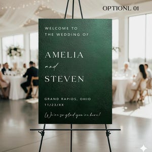 May include: A dark green wedding welcome sign on a black easel. The sign reads "WELCOME TO THE WEDDING OF AMELIA and STEVEN" with the location and date below. The text "We're so glad you're here!" is at the bottom.