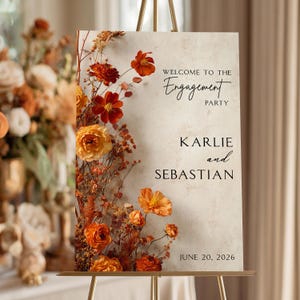May include: A rectangular engagement party sign with a textured beige background, featuring a vertical arrangement of dried orange and yellow flowers on the left side. The text reads: WELCOME TO THE Engagement PARTY, KARLIE and SEBASTIAN, JUNE 20, 2026.