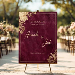 May include: A rectangular wedding welcome sign with a deep burgundy watercolor background and gold foil floral accents. The text reads: WELCOME TO OUR WEDDING, Amanda & Jack, NOVEMBER 22, 2025. The sign is displayed on a gold metal tripod easel stand.