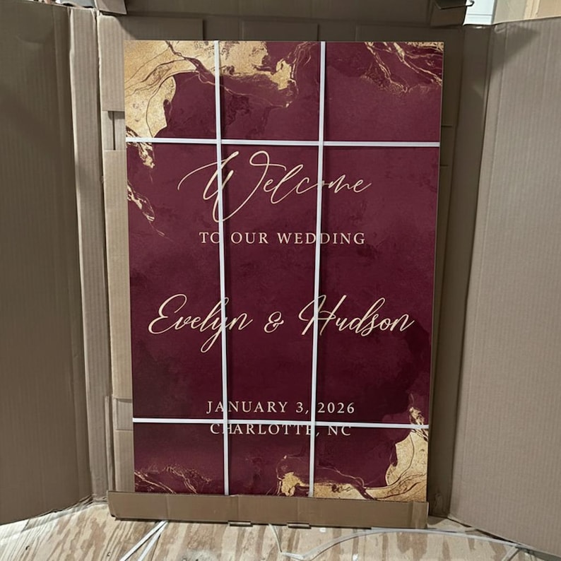 May include: A rectangular wedding welcome sign featuring a deep burgundy background with gold marble-like accents. The white text reads Welcome TO OUR WEDDING Evelyn & Hudson JANUARY 3, 2026 CHARLOTTE, NC. The sign is divided by thin white vertical and horizontal lines.