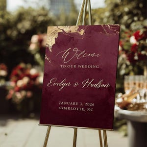 May include: A rectangular wedding welcome sign in deep burgundy with gold marble-style accents at the top. The text reads: Welcome TO OUR WEDDING Evelyn & Hudson JANUARY 3, 2026 CHARLOTTE, NC. The sign is displayed on a gold metal tripod easel.