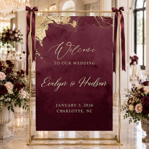 May include: A rectangular wedding welcome sign in deep burgundy with gold marble accents, suspended from a gold metal frame with dark red ribbons. The text reads: Welcome TO OUR WEDDING Evelyn & Hudson JANUARY 3, 2026 CHARLOTTE, NC.