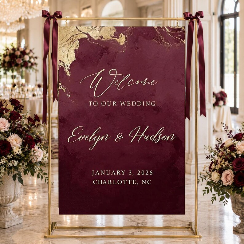 May include: A rectangular wedding welcome sign in deep burgundy with gold marble accents, suspended from a gold metal frame with dark red ribbons. The text reads: Welcome TO OUR WEDDING Evelyn & Hudson JANUARY 3, 2026 CHARLOTTE, NC.