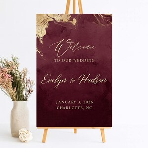 May include: A rectangular wedding welcome sign featuring a deep burgundy background with gold marble-like accents at the top. The text reads Welcome TO OUR WEDDING, Evelyn & Hudson, JANUARY 3, 2026, CHARLOTTE, NC in elegant gold script and serif fonts.