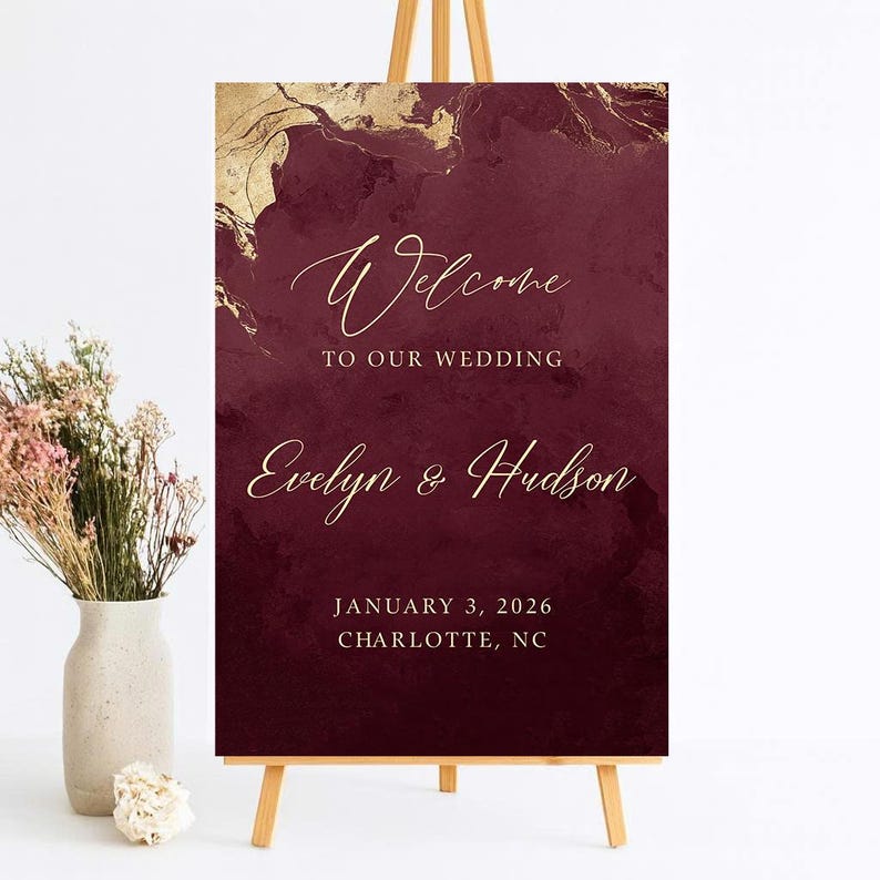 May include: A rectangular wedding welcome sign featuring a deep burgundy background with gold marble-like accents at the top. The text reads Welcome TO OUR WEDDING, Evelyn & Hudson, JANUARY 3, 2026, CHARLOTTE, NC in elegant gold script and serif fonts.