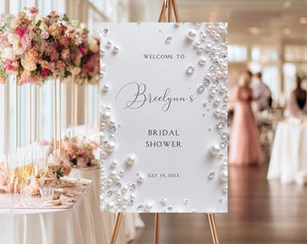 May include: A white rectangular sign on a gold easel featuring the text WELCOME TO Breelynn's BRIDAL SHOWER JULY 19, 20XX. The sign is decorated with a border of white pearls and clear sparkling crystals arranged along the left and right edges.