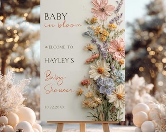 May include: A rectangular sign on a wooden easel featuring the text BABY in bloom WELCOME TO HAYLEY'S Baby Shower 10.22.20XX. The right side displays a vertical arrangement of textured, painted flowers in shades of pink, yellow, blue, and white.