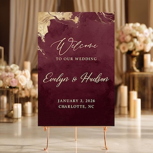 May include: A rectangular wedding welcome sign featuring a deep burgundy background with gold marble-like accents at the top. The text reads: Welcome TO OUR WEDDING Evelyn & Hudson JANUARY 3, 2026 CHARLOTTE, NC in elegant gold script and block lettering.