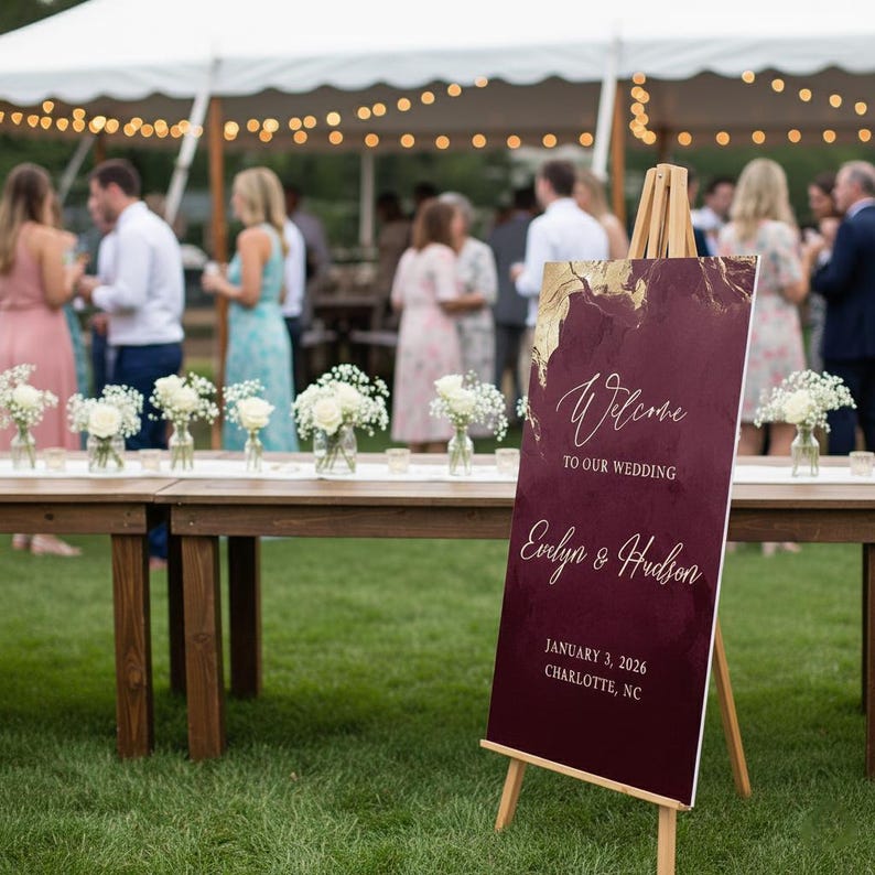May include: A vertical rectangular wedding welcome sign on a wooden easel, featuring a deep burgundy background with gold marble-like accents. The text reads: Welcome to our wedding, Evelyn & Hudson, January 3, 2026, Charlotte, NC in white script.