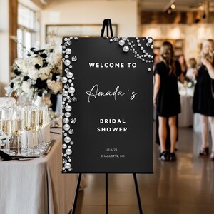 May include: Black sign with white text that reads "Welcome to Amada's Bridal Shower" with date and location. The sign is decorated with pearl and rhinestone accents and is displayed on a black easel.