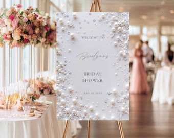 Pode incluir: Uma placa de boas-vindas retangular branca num cavalete dourado, decorada com pérolas brancas e cristais transparentes espalhados pelas bordas. O texto diz: Welcome to Breelynn's Bridal Shower July 19, 20XX, com letras pretas.