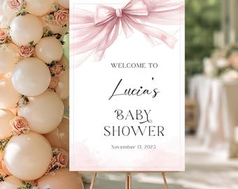 May include: A rectangular welcome sign featuring a watercolor illustration of a large pink bow at the top. The text reads: WELCOME TO Lucia's BABY SHOWER November 13, 2025. The sign is displayed on a wooden easel next to a balloon and floral arch.