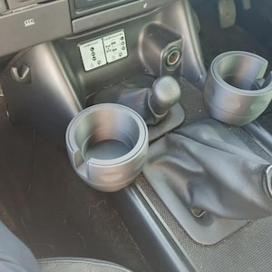 Könnte beinhalten: Drei graue Kunststoff-Getränkehalter sind im Inneren eines Autos abgebildet. Zwei sind eingebaut, einer ist lose. Die Getränkehalter sind für die Mittelkonsole des Autos konzipiert. Der Schalthebel und andere Bedienelemente des Autos sind ebenfalls sichtbar.