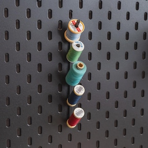 May include: A collection of colorful thread spools, including red, blue, teal, and green, are arranged on a dark gray pegboard. The spools are various sizes and have white labels. The pegboard has a pattern of evenly spaced, oblong holes.