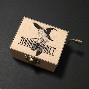 Op de afbeelding: Een houten muziekdoos met de tekst "TOUHOU PROJECT" en een zwarte illustratie van een heks op het deksel. De doos heeft een metalen slinger en een messing sluiting. De doos is gemaakt van lichtgekleurd hout.