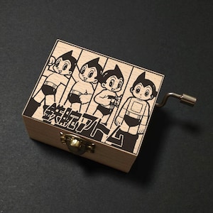May include: A wooden music box featuring a black and white illustration of Astro Boy characters. The box has a brass clasp and a silver crank. The Japanese text is visible on the front of the box.