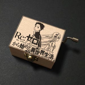 Può includere: Carillon in legno con illustrazione anime in bianco e nero. Il coperchio presenta testo giapponese e il titolo "Re:Zero". Una manovella argentata è attaccata al lato. La scatola è chiusa con un fermaglio color ottone.