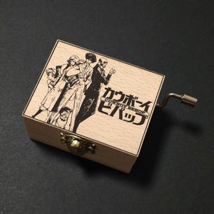 Può includere: Carillon in legno con un'illustrazione in bianco e nero di personaggi e testo giapponese. Ha una manovella in metallo e una chiusura in ottone. Il design è ispirato alla serie anime Cowboy Bebop.