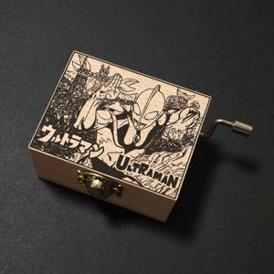 May include: Wooden music box featuring a laser-etched design of Ultraman, with Japanese text and the English word "Ultraman." The box has a brass clasp and a silver crank. The artwork depicts Ultraman in a dynamic pose.