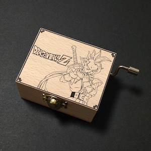 Puede incluir: Caja de música de madera con un diseño grabado con láser de un personaje de Dragon Ball Z. La caja tiene una tapa con bisagras, un cierre metálico y una manivela. El texto "DRAGONBALL Z" está grabado en la tapa.