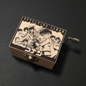 May include: A wooden music box featuring a black and white illustration of characters from the anime series "Detective Conan." The box has a hinged lid, a metal crank, and a brass-colored clasp. The text "Detective Conan" is printed on the top.