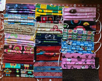 May include: A collection of 15 colorful fabric face masks with various patterns and designs. The masks are arranged in rows on a brown surface with a white ruler in the background. Some of the patterns include a galaxy print, a basketball print, a floral print, a geometric print, and a cartoon print. The masks are all made of cotton fabric and have elastic ear loops.
