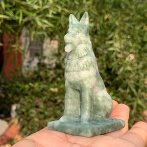 May include: A green and white stone sculpture of a dog, possibly a German Shepherd, sitting upright on a square base. The dog has pointed ears and a detailed face. The sculpture is held in a person's hand, with a blurred green background.