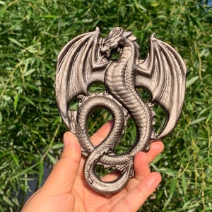 May include: A detailed, metallic dragon sculpture with outstretched wings and a serpentine body. The dragon is a bronze color and is held against a backdrop of green foliage. The sculpture is a decorative item.