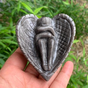 May include: A grey stone sculpture of two figures embracing within a heart-shaped frame. The sculpture has a textured surface and is held in a hand against a backdrop of green foliage. The figures are in a seated position.