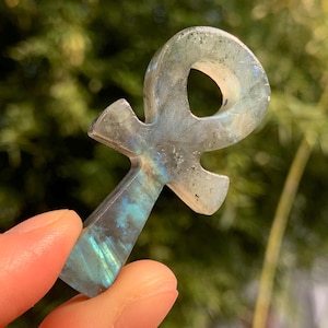 May include: A carved ankh pendant made of iridescent labradorite. The ankh, a symbol of life, features a cross shape with a loop at the top. The stone displays shades of blue, green, and gray, with a polished finish. The background is a blurred view of green foliage.