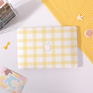 May include: A laptop with a yellow and white gingham pattern case, featuring the Apple logo. The case is on a white surface, with a yellow gingham cloth and small decorative items nearby. The overall theme is a cheerful, summery aesthetic.