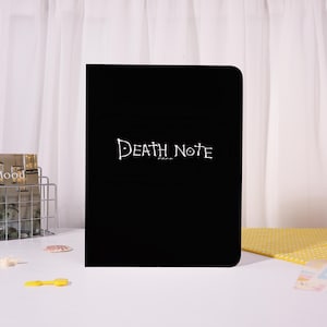 May include: A black notebook with rounded corners. The title "Death Note" is written in white script across the front. The notebook is sitting on a white surface.