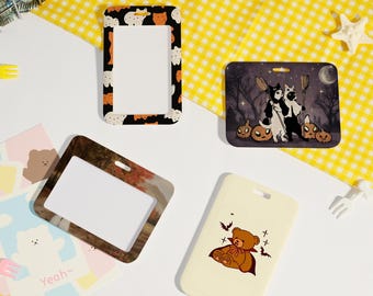 May include: A collection of decorative ID badge holders with various designs. One holder features a black frame with orange and white cat faces. Another has a Halloween-themed illustration of cats and pumpkins. A third has a bear with a cape.