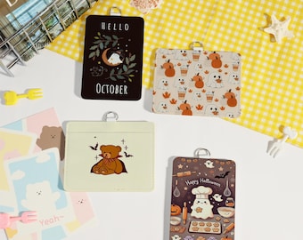 May include: A collection of cardholders with various designs. One cardholder is black with "Hello October" text and a moon and ghost graphic. Other cardholders feature pumpkins, ghosts, and a teddy bear. A "Happy Halloween" cardholder shows a ghost chef.