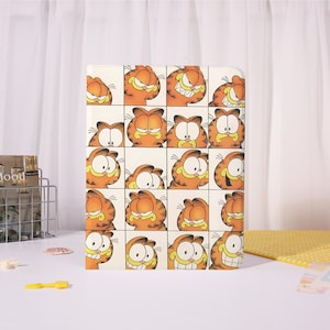 May include: A rectangular, cream-colored tablet case featuring a grid of Garfield the cat's face in various expressions. The cartoon cat is orange with black stripes and a yellow background.