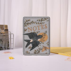 May include: A rectangular notebook with a gray background and the word "NOTES" in orange. The cover features a dark gray bird in flight, a yellow sun, and a moth. Decorative leaves and branches surround the edges.