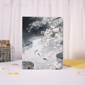 May include: A notebook with a serene watercolor painting of a pond with koi fish and cherry blossoms. The artwork features shades of gray, white, and touches of red and orange. The cover has a glossy finish.