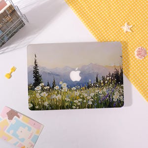 May include: A laptop hard shell case featuring a printed landscape illustration of a mountain range behind a field of white and yellow wildflowers and tall pine trees, with a central cutout revealing the silver Apple logo on the laptop lid.