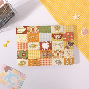 May include: A laptop skin featuring a grid pattern of various illustrations including yellow bows, red hearts with a bear face, bouquets of flowers, green hearts, and checkered or polka dot squares in shades of yellow, orange, green, and white.