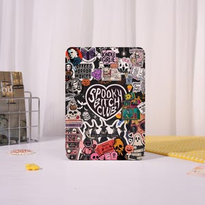 May include: A black Kindle case covered in stickers with a spooky theme. The stickers include images of skulls, ghosts, pumpkins, and text that reads "Spooky Bitch Club", "Coffee & Horror Movies", and "Kindle Girlie".