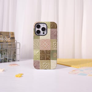 Puede incluir: Una funda rectangular para smartphone con una cuadrícula de azulejos cuadrados en relieve en tonos apagados de verde salvia, rosa empolvado, crema y bronce. Cada azulejo muestra un patrón floral o geométrico en relieve único.