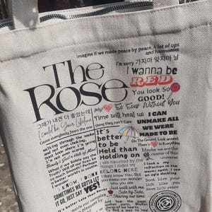 Może przedstawiać: Pł&oacute;cienna torba w kolorze złamanej bieli z czarnym tekstem i grafiką. Tekst zawiera "The Rose" i teksty piosenek w języku angielskim i koreańskim. Posiada zapięcie na zamek błyskawiczny i uchwyty z tkaniny.
