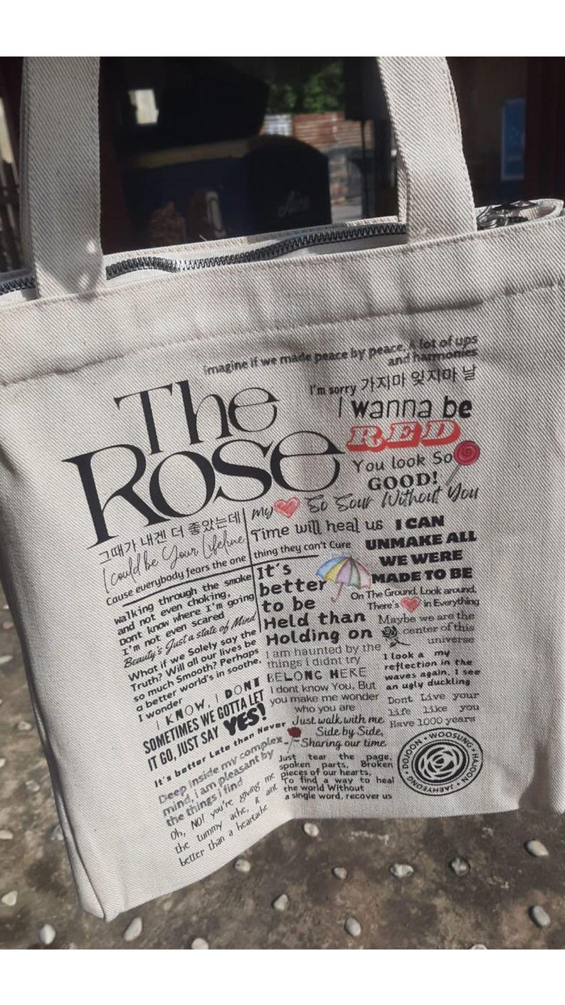 Może przedstawiać: Pł&oacute;cienna torba w kolorze złamanej bieli z czarnym tekstem i grafiką. Tekst zawiera "The Rose" i teksty piosenek w języku angielskim i koreańskim. Posiada zapięcie na zamek błyskawiczny i uchwyty z tkaniny.