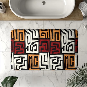 May include: Rectangular bath mat with a geometric pattern in black, white, red, and orange. The design features abstract shapes and lines, creating a bold and colorful statement. The mat is placed in front of a white bathtub.