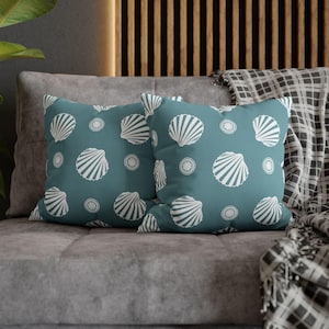 Op de afbeelding: Twee turquoise kussens met een wit schelp- en cirkelpatroon. Ze liggen op een grijze bank, met een patroon deken op de achtergrond. Decoratieve woondecoratie.