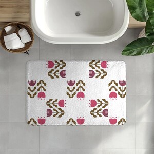 Puede incluir: Alfombra de baño rectangular blanca con un patrón repetido de flores rosas en forma de tulipán y tallos marrones. La alfombra está colocada frente a una bañera blanca, con una cesta de toallas cerca. El tema es la decoración floral del baño.