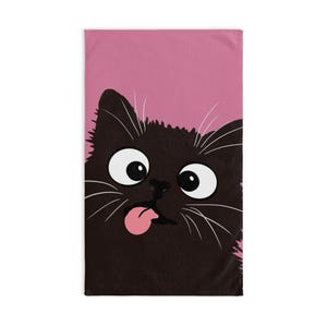 May include: A rectangular towel featuring a graphic illustration of a black cat with large white eyes and a pink tongue sticking out. The cat is positioned against a solid pink background, with long white whiskers extending from its face.