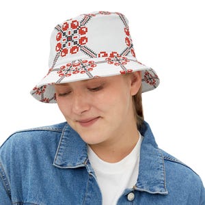 Puede incluir: Sombrero de pescador blanco con un patrón repetido de diseños florales geométricos rojos, negros y blancos. El sombrero tiene un ala ancha y una corona suave. La persona lleva una chaqueta vaquera.